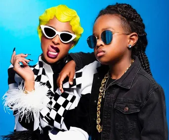 Two individuals in stylish costumes, one with a yellow wig and sunglasses, both wearing chic black and white outfits on a blue background.