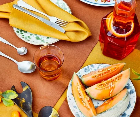 Colorful table setting with orange napkin, cutlery, glass of amber drink, cantaloupe slices, and carafe with red liquid and fruit.