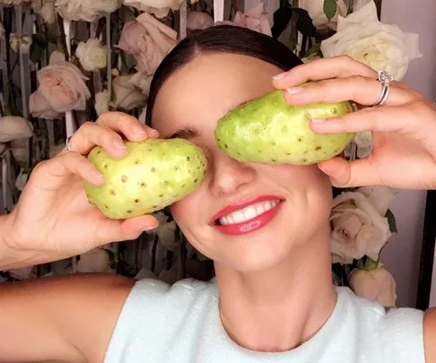 Woman smiling and playfully holding two noni fruits over her eyes, with a floral background.