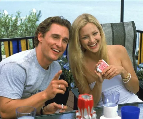 A man and a woman smiling outdoors, seated at a table with drinks and playing cards, enjoying a sunny day.