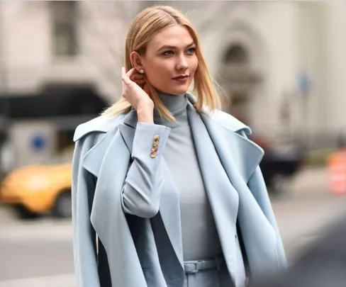 Woman in light blue coat and turtleneck, touching her hair while walking in a city street.