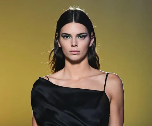 A model in a black dress on a runway with green eye makeup against a yellow background.