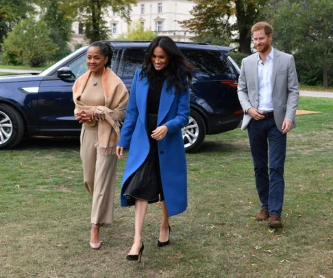 Meghan Markle, Prince Harry, and Doria Ragland walking outdoors near a car, with trees and a building in the background.