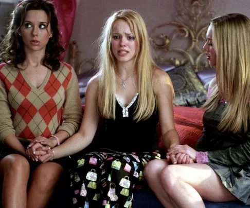 Three girls sit on a bed holding hands; middle girl looks emotional. Scene from "Mean Girls."