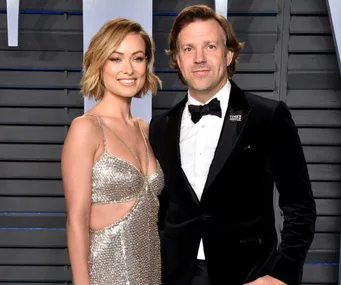 A man and woman dressed in formal attire pose together, smiling, in front of a gray background at an event.