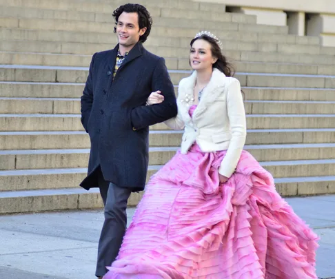 A woman in a white jacket and pink gown with a tiara walks arm-in-arm with a man in a dark coat on steps.