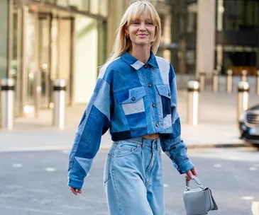 Woman in patchwork denim jacket and jeans, holding a small handbag, walking on city street.