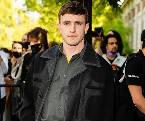 Man in a dark jacket standing outdoors at an event, with people in the background.