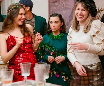 Three women in festive attire laughing at a party; one in a red dress, another in green, and the third in white.