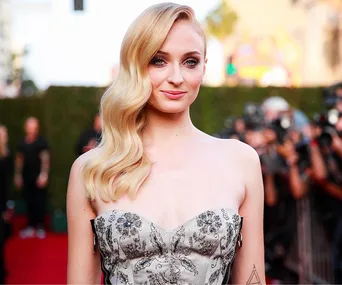 A woman with long blonde hair, wearing a strapless dress, poses on a red carpet with photographers in the background.