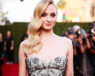 A woman with long blonde hair, wearing a strapless dress, poses on a red carpet with photographers in the background.