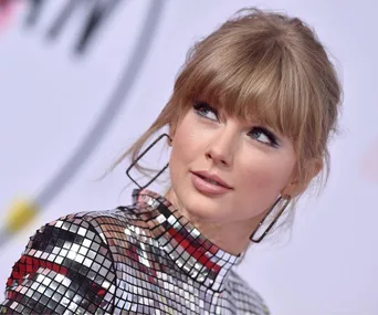 A person with blonde hair looks upward, wearing a shiny, metallic outfit with large hoop earrings.