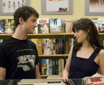 Two people smiling at each other in a record store. Shelves filled with DVDs and music can be seen behind them.