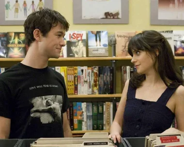 Two people smiling at each other in a record store. Shelves filled with DVDs and music can be seen behind them.