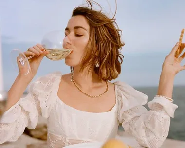 Woman in white dress sips wine outdoors, holding breadstick; ocean background.