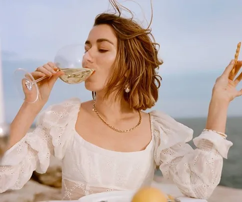 Woman in white dress sips wine outdoors, holding breadstick; ocean background.