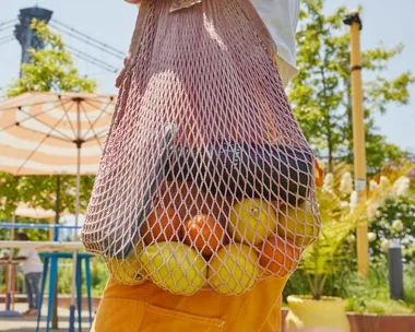 Mesh bag with apples, oranges, and a bottle, carried by a person outdoors near patio seating and plants.