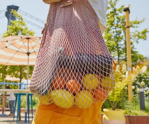 Mesh bag with apples, oranges, and a bottle, carried by a person outdoors near patio seating and plants.