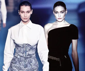 Two models on a runway, one in a white blouse and corset, the other in a black one-shoulder dress, with slicked-back hair.