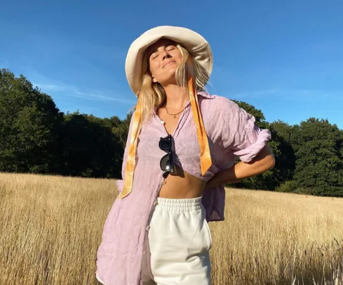 Woman in a lavender shirt and white shorts with a broad hat stands in a field under clear skies.