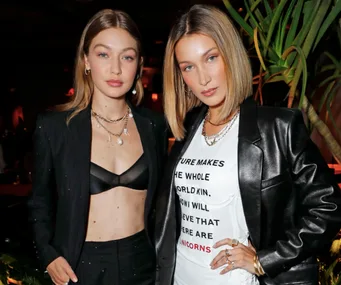 Two women posing stylishly in a dimly lit setting, one in a black suit and the other in a leather jacket.