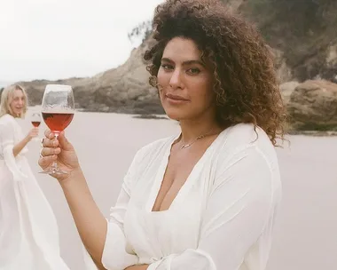 Woman in white dress holding a glass of red wine on a beach, with another woman in background smiling.