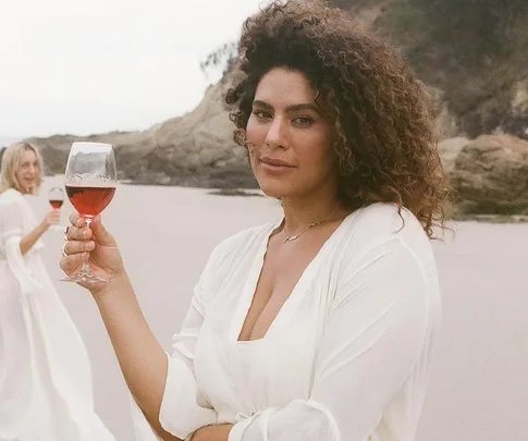 Woman in white dress holding a glass of red wine on a beach, with another woman in background smiling.