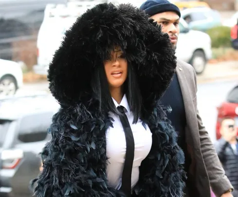 Woman in a black feathered coat with a hood, white shirt, and black tie, walking outside with a crowd nearby.