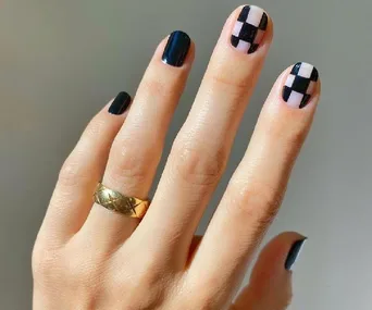 A hand with a gold ring features checkerboard nail art in black and nude colors on the nails.