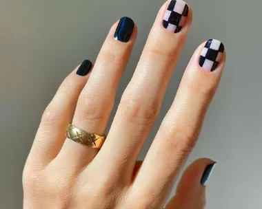 A hand with a gold ring features checkerboard nail art in black and nude colors on the nails.