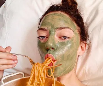 A person with a green facial mask eating spaghetti in bed.