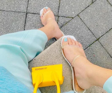 Feet in clear sandals with blue nail polish, wearing blue pants, next to a small yellow "JACQUEMUS" handbag on pavement.