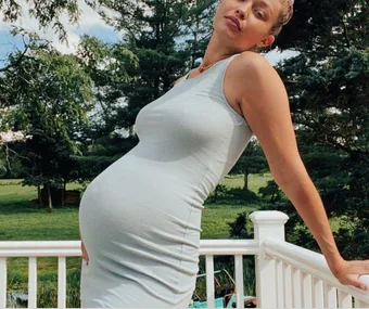 Pregnant woman in a fitted dress poses outdoors on a sunny day with trees in the background.