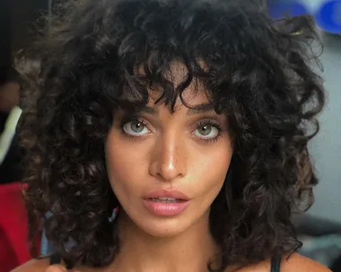 Woman with curly dark hair and bangs looking directly at the camera, wearing a sleeveless top.