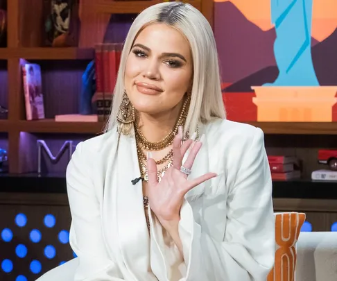 A woman with long blonde hair smiles, wearing a white outfit and jewelry, sitting in a room with a colorful background.