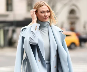 A woman in a stylish light blue coat and turtleneck walking on a city street, with blurred taxis in the background.