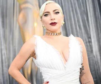 Lady Gaga in a white dress with blonde hair, wearing a bold necklace, standing against a silver curtain backdrop.