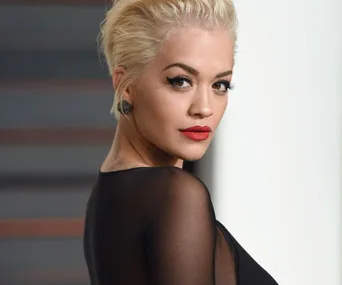 A woman with short blonde hair and red lipstick, wearing a black outfit, looks over her shoulder.