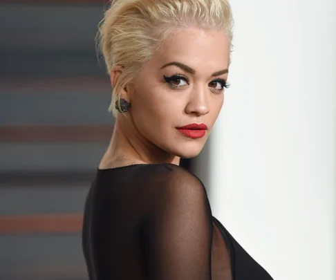 A woman with short blonde hair and red lipstick, wearing a black outfit, looks over her shoulder.