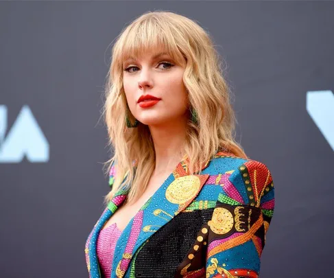 A woman with wavy blonde hair in a colorful patterned jacket poses against a dark background.