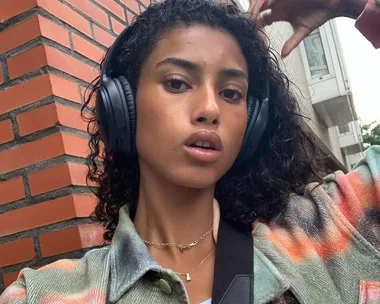 Young woman with curly hair wearing headphones, patterned jacket, standing outside near a brick wall, looking at the camera.