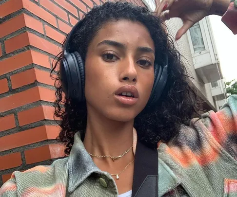 Young woman with curly hair wearing headphones, patterned jacket, standing outside near a brick wall, looking at the camera.