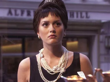 Woman in a black dress with pearls and a tiara gazes out a shop window, reminiscent of a classic film scene.