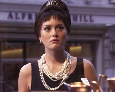 Woman in a black dress with pearls and a tiara gazes out a shop window, reminiscent of a classic film scene.