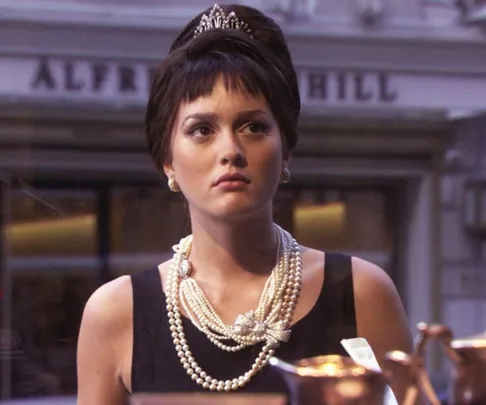 Woman in a black dress with pearls and a tiara gazes out a shop window, reminiscent of a classic film scene.