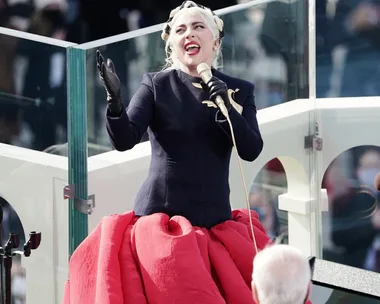 Lady Gaga singing at the 2021 inauguration, wearing a black top and red skirt with a gold dove brooch.