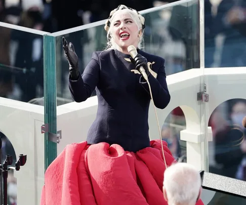 Lady Gaga singing at the 2021 inauguration, wearing a black top and red skirt with a gold dove brooch.