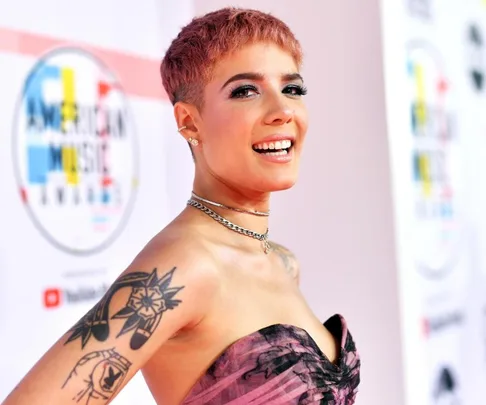 Singer with short hair and tattoos smiling on a red carpet, American Music Awards backdrop in the background.