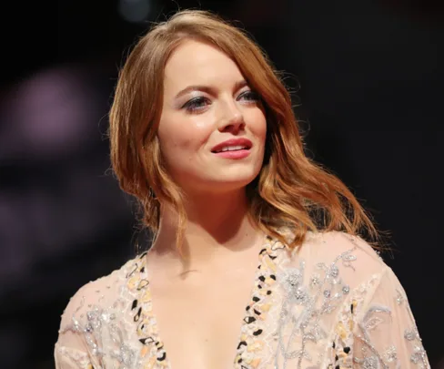 Red-haired woman in a detailed embroidered dress stands under soft lighting, looking slightly off-camera with a faint smile.