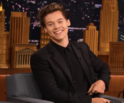 Smiling person in a black suit seated on a talk show set with a cityscape backdrop.